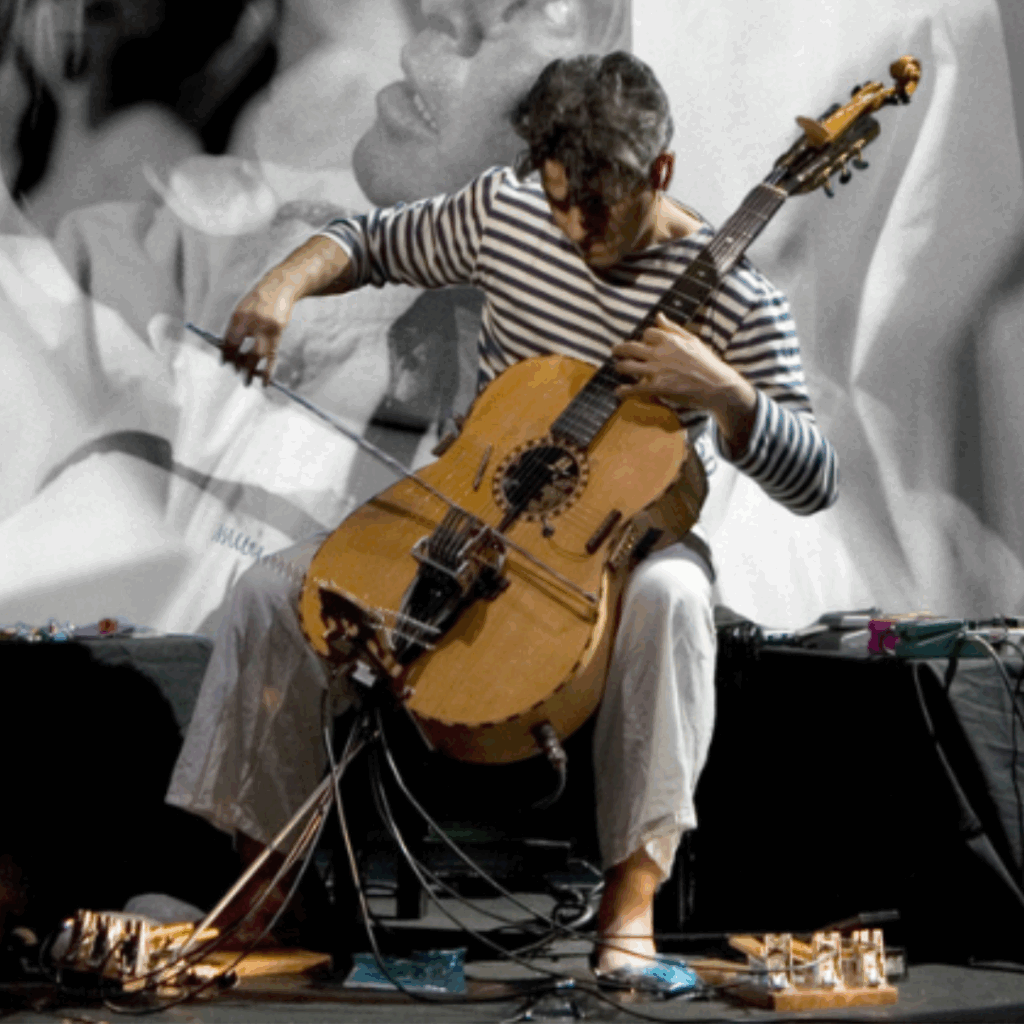 Paolo Angeli plays his prepared Sardinian guitar on stage, with a large black-and-white projection of two faces behind him.