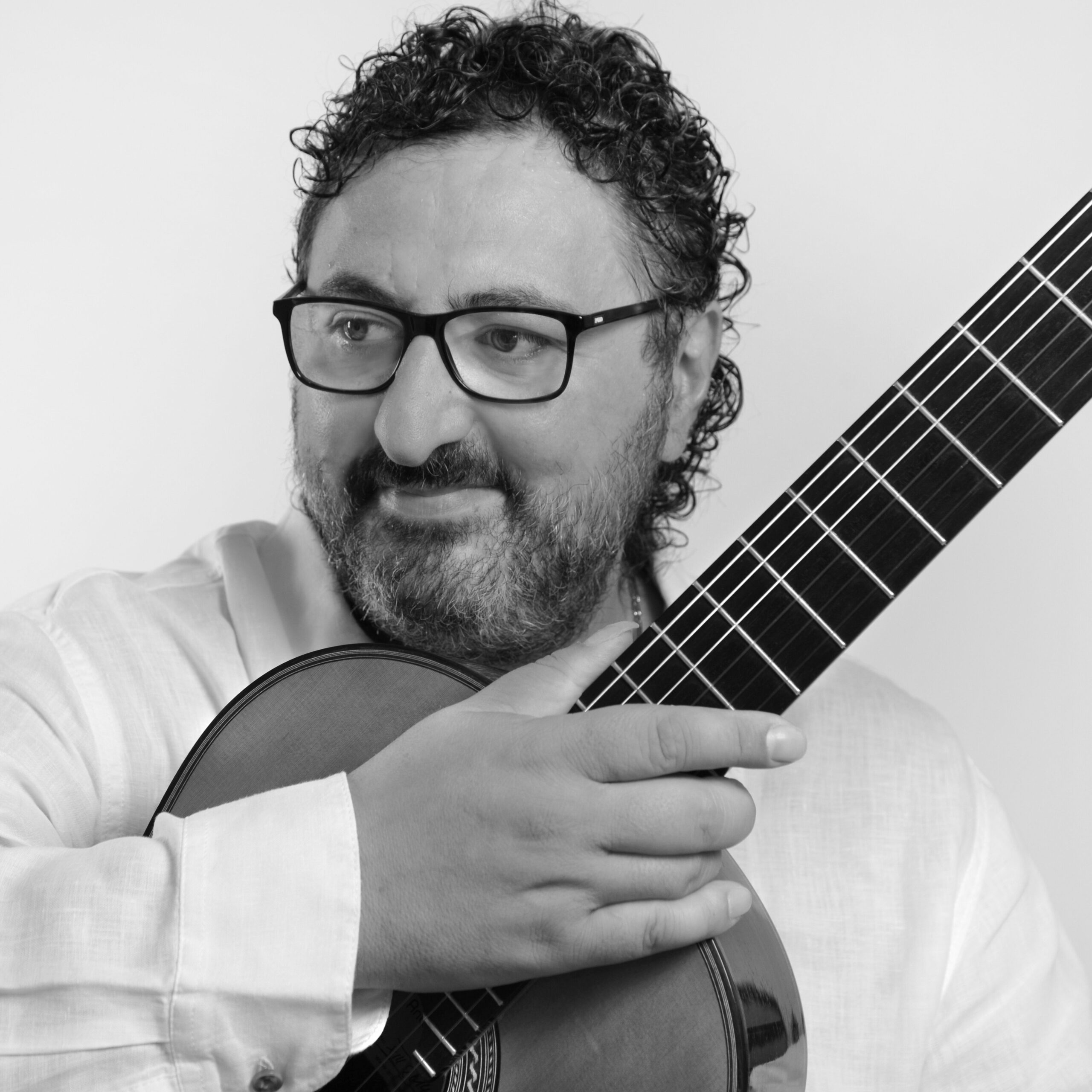 Black-and-white portrait of a bearded guitarist wearing glasses and holding a classical guitar close to his chest.