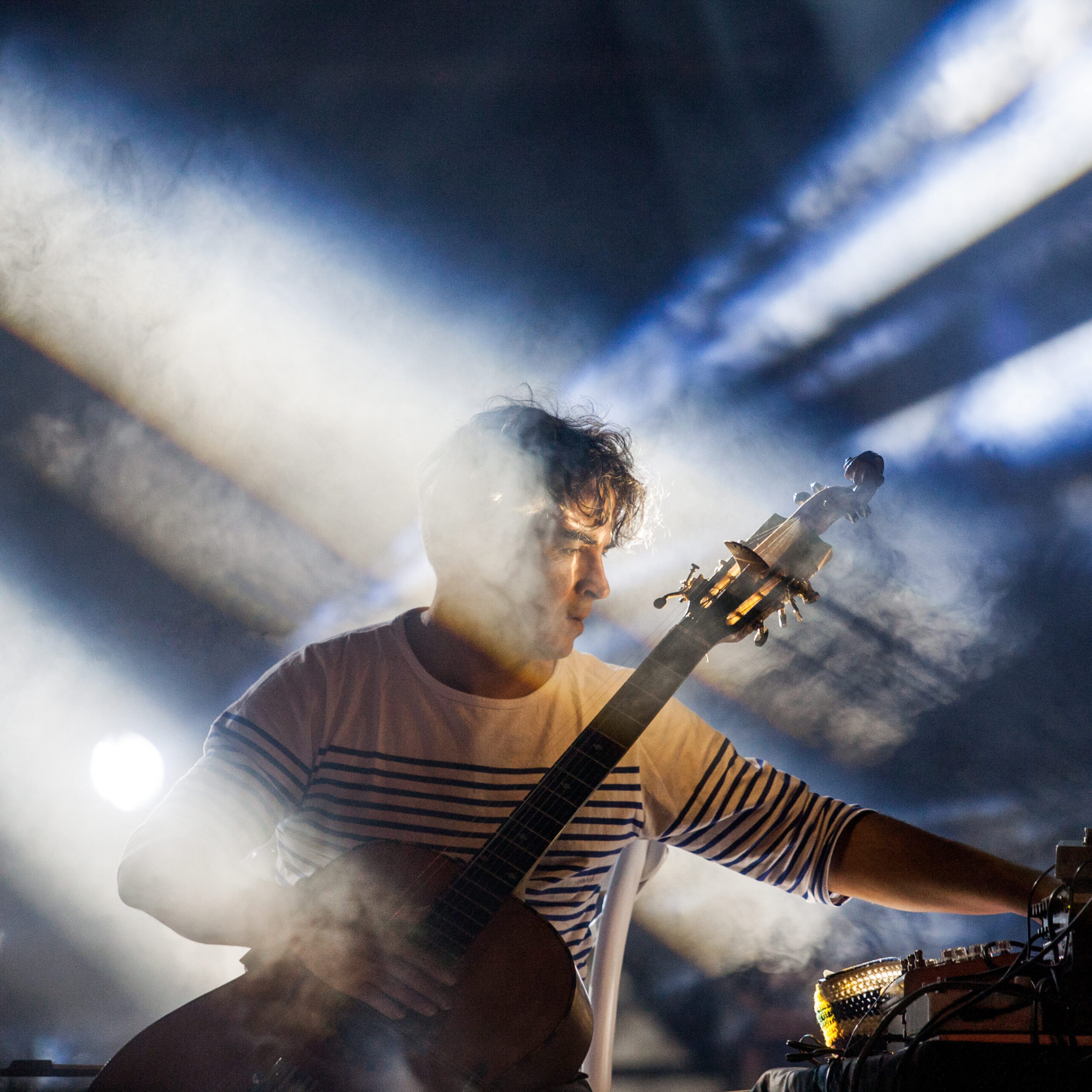 A guitarist performs onstage amid dramatic beams of light and smoke, reaching toward electronic equipment while holding a guitar.