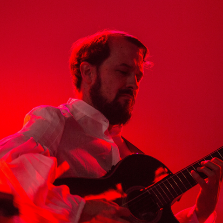 A bearded guitarist performs onstage under dramatic red lighting, playing a classical guitar.