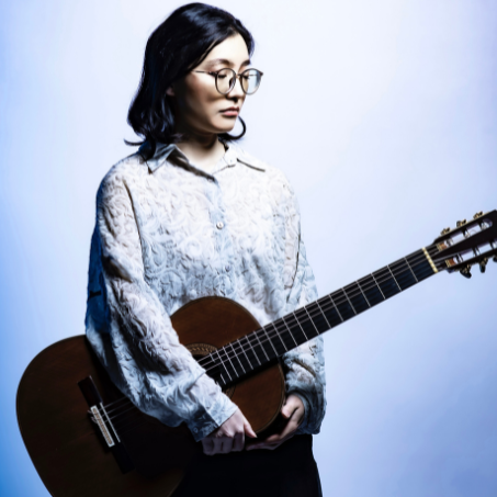 A woman wearing glasses holds a classical guitar against a softly lit blue-and-white background.