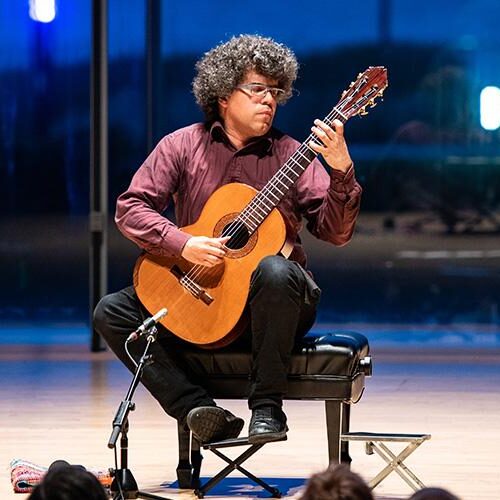 A classical guitarist performs seated onstage, playing guitar in a modern concert hall.